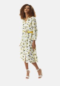 White dress with yellow floral print, buttoned front, and long sleeves, cinched at the waist with a yellow belt. Worn with white strappy heels.