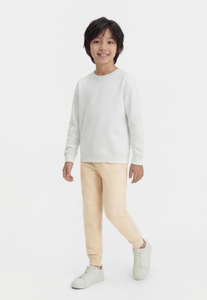 Ragazzo in piedi che sorride, indossa una felpa bianca, pantaloni jogger beige e sneakers bianche, su sfondo chiaro semplice.