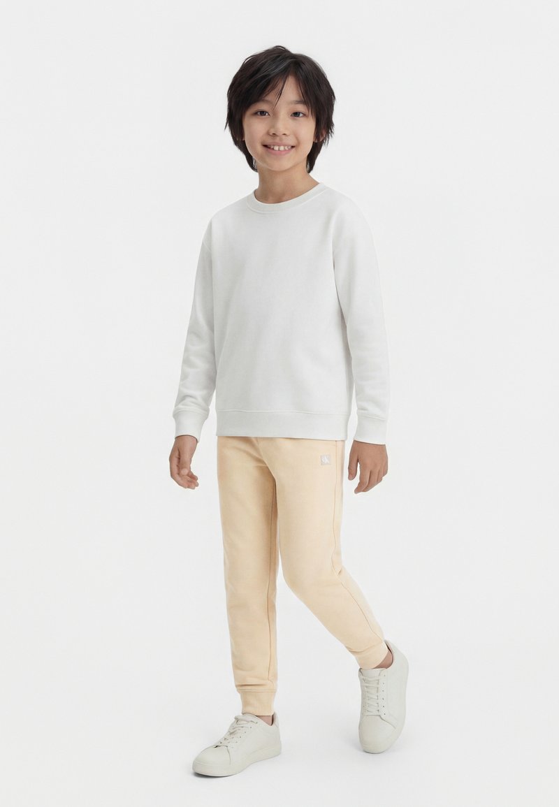 Αγόρι που στέκεται και χαμογελά, φορώντας λευκό φούτερ, μπεζ παντελόνι τύπου jogger και λευκά αθλητικά παπούτσια, με φόντο ένα απλό ανοιχτόχρωμο.