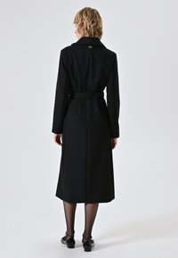 Persona in piedi di spalle che indossa un lungo cappotto nero con cintura, collant neri trasparenti e scarpe nere con tacco basso su uno sfondo neutro.