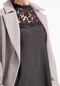 Manteau gris avec un col structuré, superposé à un haut noir présentant un décolleté en dentelle florale. Tissu lisse avec une finition mate.