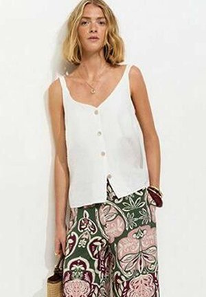 Femme portant un top blanc sans manches boutonné et un pantalon ample à motifs verts, roses et blancs, debout contre un mur blanc.