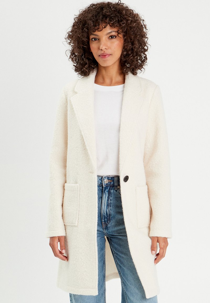 Manteau en mélange de laine texturé blanc avec col cranté, fermeture à un bouton et deux poches avant, associé à un t-shirt blanc et un jean en denim.