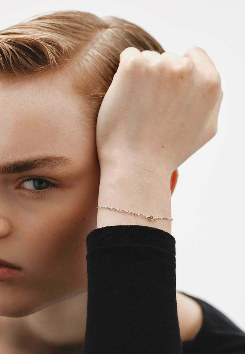 Una pulsera de plata en una muñeca presenta una delicada cadena y un pequeño charm circular. El fondo es neutro, destacando la joyería.