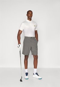 Golf outfit featuring a white polo shirt with yellow logo, gray shorts, and white golf shoes with blue accents. Holding a golf club.