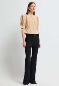 Vrouw in een beige blouse met pofmouwen en knopen, met zwarte hoge taille flare broeken en zwarte puntige schoenen, staand tegen een effen achtergrond.
