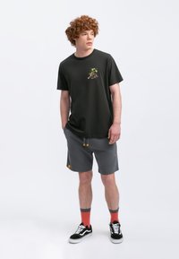 Zwarte katoenen T-shirt met een kleine kleurrijke graphic, gecombineerd met grijze sportshorts met gele accenten en zijzakken. Zwarte sneakers en rode sokken.