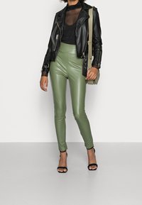 Mujer que lleva leggings de cuero verde de tiro alto, blusa negra transparente, chaqueta de cuero negra, tacones negros y lleva un bolso de hombro Beige.