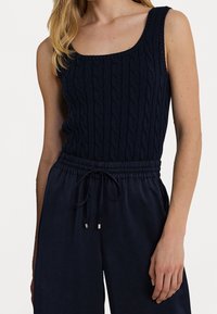 Marinblå stickad linne med flätmönster, med fyrkantig halsringning och breda axelband. Matchas med mörka byxor med dragsko.