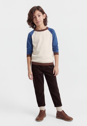 Αγόρι με μαλλιά μέχρι τον ώμο, ντυμένο με κρεμ και μπλε μακρυμάνικο πουκάμισο, σκοτεινό παντελόνι, κρεμ κάλτσες και καφέ παπούτσια, στέκεται χαλαρά σε λευκό φόντο.