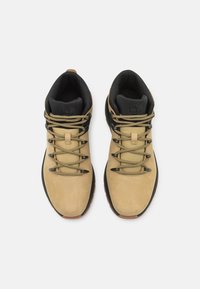 Scarpe da trekking in suede tan con dettagli neri, lacci rotondi e un collare di supporto. Suola in gomma testurizzata per aderenza. Logo visibile sulla linguetta.