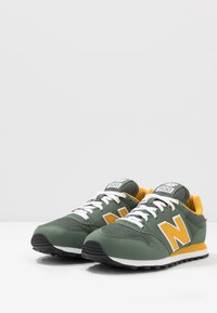Sneaker verdi con dettagli gialli, mesh testurizzato e materiale sintetico, lacci bianchi e suola in gomma nera, con un grande logo sul lato.