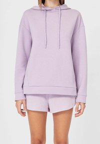 Mujer con una sudadera ligera de color púrpura con cordones y pantalones cortos a juego, de pie frente a un fondo blanco liso.