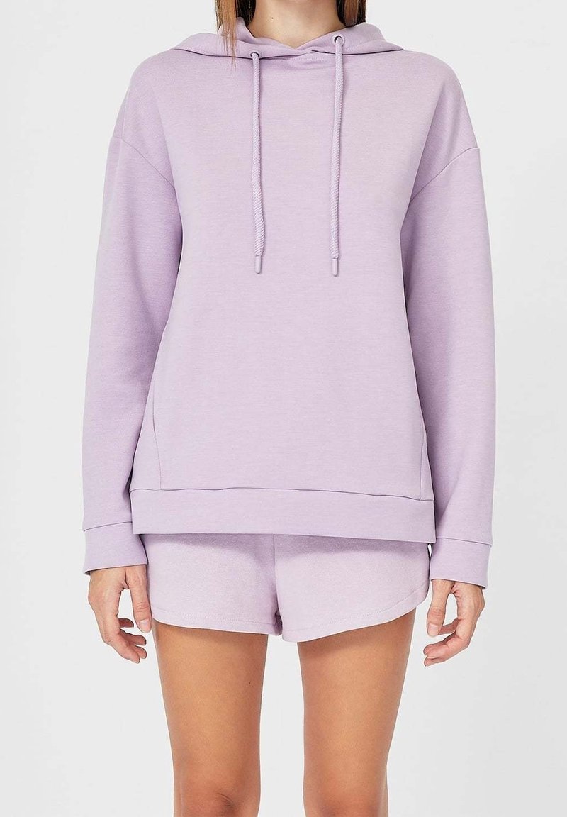 Mujer con una sudadera ligera de color púrpura con cordones y pantalones cortos a juego, de pie frente a un fondo blanco liso.
