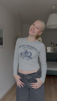 Leende kvinna med blont hår i en hästsvans bär en grå crop top med långa ärmar och mörka jeans, står i ett ljust sovrum.