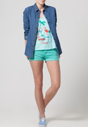 Chemise en denim à boutons, short vert menthe et baskets bleu clair. Débardeur graphique mettant en avant des flamants roses et des palmiers aux couleurs vives.