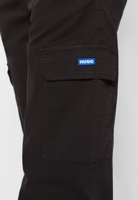 Czarne spodnie cargo z przednią kieszenią; posiadają niebieską łatkę z logo "HUGO". Wykonane z trwałego materiału, o gładkiej fakturze.