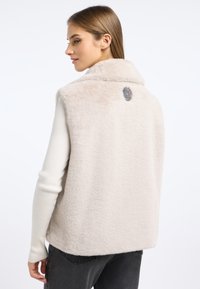 Beżowa kamizelka ze sztucznego futra z kołnierzem, z metalowym akcentem w formie medalu z tyłu. Miękka tekstura, bez rękawów i z bocznymi szwami.