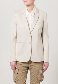 Beige sako s přiléhavým střihem, s klopovými límci, na dva knoflíky a bočními kapsami. Nosené přes bílé tričko, doplněné khaki kalhotami.