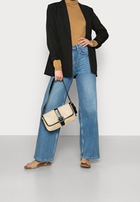 Blazer noir sur un col roulé beige, jeans à taille haute et coupe large bleu clair, et sandales beiges. Un sac en osier avec des accents noirs est tenu.