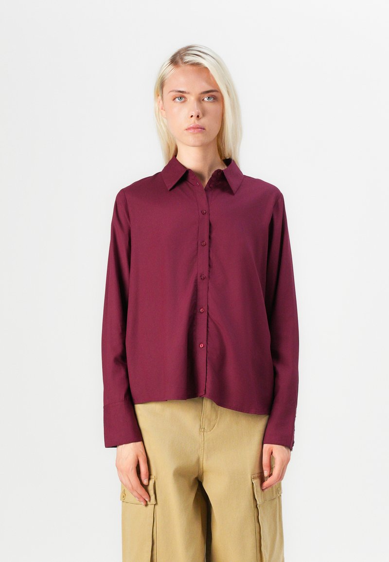 Burgundy long-sleeve button-up shirt with a pointed collar, smooth texture, and a straight hem, paired with beige cargo pants.