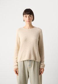 Beige színű, strukturált pulóver hosszú ujjakkal és kerek nyakkivágással, világoskék, laza fazonú nadrággal párosítva, sima háttér előtt bemutatva.