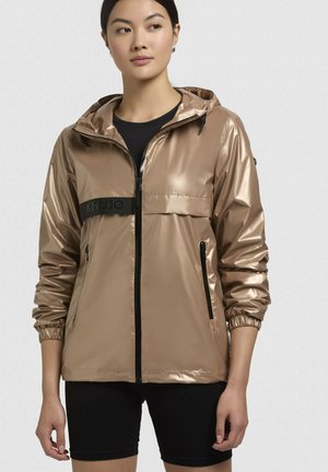 Metalická béžová bunda s kapucňou, so zapínaním na zips, čiernymi akcentmi, elastickými manžetami a logovým pásom. Hladká, lesklá textúra.