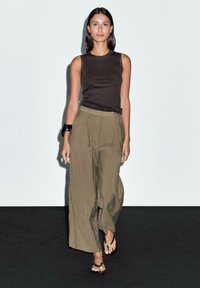 Top nero senza maniche, pantaloni verde oliva a vita alta e larghi, bracciale nero di grandi dimensioni e infradito nere su uno sfondo neutro.