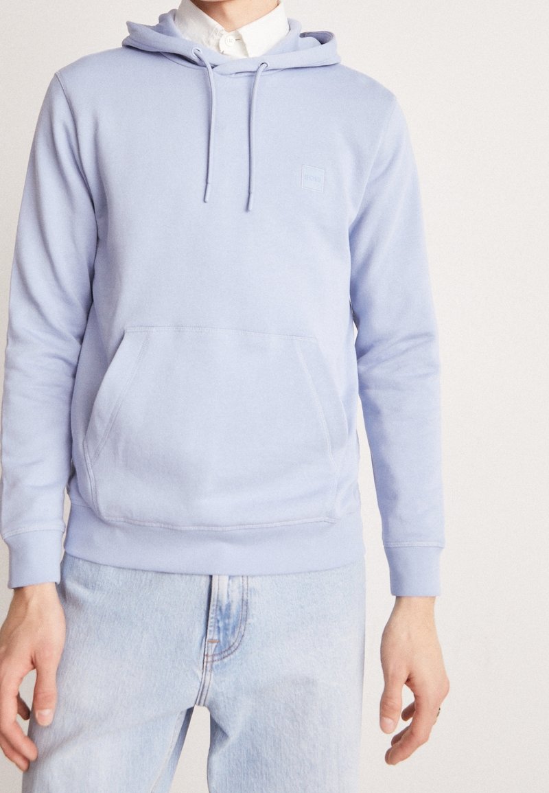 Personne portant un sweat à capuche bleu clair avec poche avant sur une chemise blanche à col et un jean bleu clair sur un fond uni.