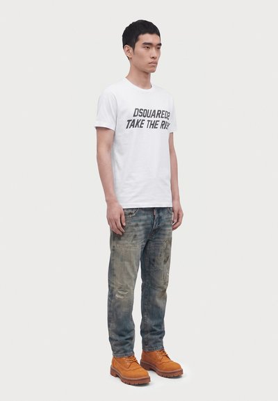 Jeune homme portant un T-shirt blanc avec le texte "DSQUARED2 TAKE THE RISK", un jean délavé et des bottes de travail orange-brun, debout de profil.