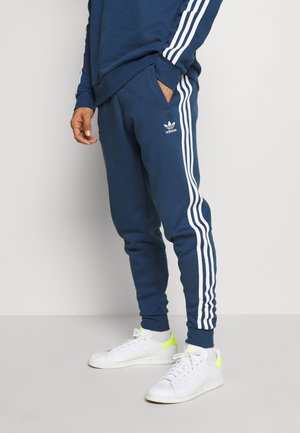 Mężczyzna ubrany w granatowe dresy Adidas i bluzę z kapturem z białymi paskami, w zestawie z białymi sneakersami z neonowo-żółtymi akcentami.