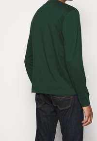 Mörkgrön långärmad t-shirt i bomull med rund hals, med en slät yta. Matchad med mörka jeans i denim, som har kontrasterande sömmar.