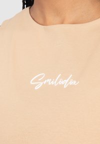 Světlé béžové tričko s hladkou texturou. Na hrudní části se nachází bílý kuršívní logo 'Smilodon'. Klasický design s kulatým výstřihem.