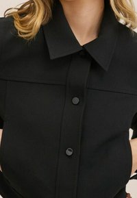 Chemise noire à col avec une patte de boutonnage ornée de boutons ronds, un design structuré et une texture de tissu lisse.