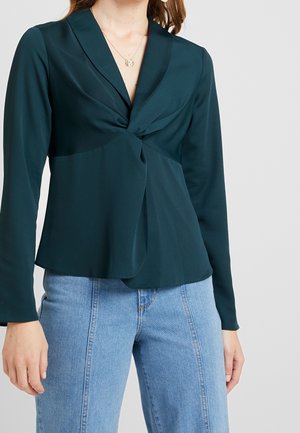 Vrouw draagt een donkergroene blouse met lange mouwen en een gedraaid voorpanddetail en lichtblauwe high-waisted jeans.