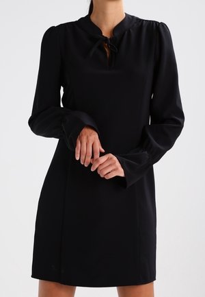 Femme portant une robe noire à manches longues avec des épaules bouffantes et un détail noué au col, debout les mains doucement jointes.