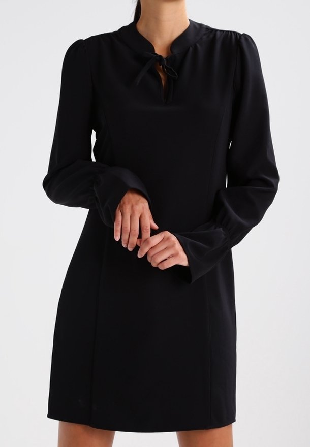 Femme portant une robe noire à manches longues avec des épaules bouffantes et un détail noué au col, debout les mains doucement jointes.