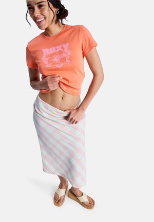 Femme souriante levant un t-shirt orange Roxy, portant une jupe midi à rayures pastel, des sandales compensées beige et un bandeau à motifs.