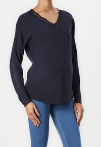 Kvinna klädd i en mörk marinblå V-ringad tröja med spetsdetaljer, ihop med ljusblå åtsittande jeans, mot en vit bakgrund.