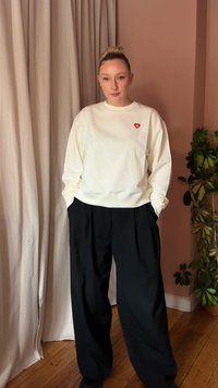 Junge Person mit blonden Haaren im Dutt trägt ein weißes Sweatshirt mit einem kleinen roten Herz-Emblem und lockere schwarze Hosen, steht drinnen.