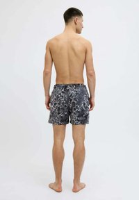 Uomo in piedi a piedi nudi, girato di spalle, che indossa pantaloni da bagno a motivo d'acqua neri e grigi con una tasca sul retro, su uno sfondo grigio chiaro.