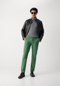 Sort læderjakke, grå turtleneck sweater, grønne bukser og sorte sko. Model iført solbriller, står op ad en ensfarvet baggrund.