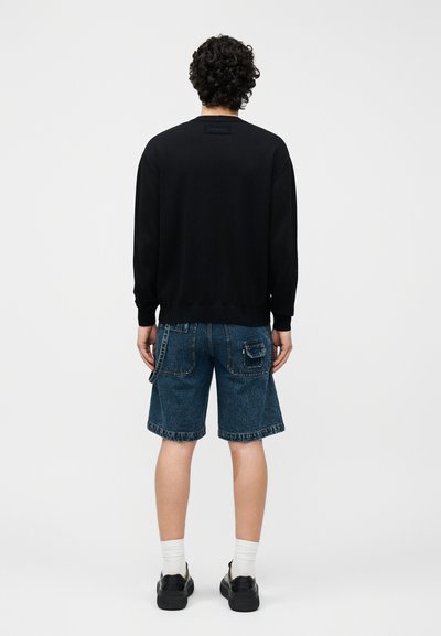 Άνδρας με σγουρά μαλλιά που φοράει μαύρο μακρυμάνικο πουκάμισο, μπλε τζιν σορτς με τσέπες, λευκές κάλτσες και μαύρα παπούτσια, στέκεται γυρισμένος με την πλάτη.