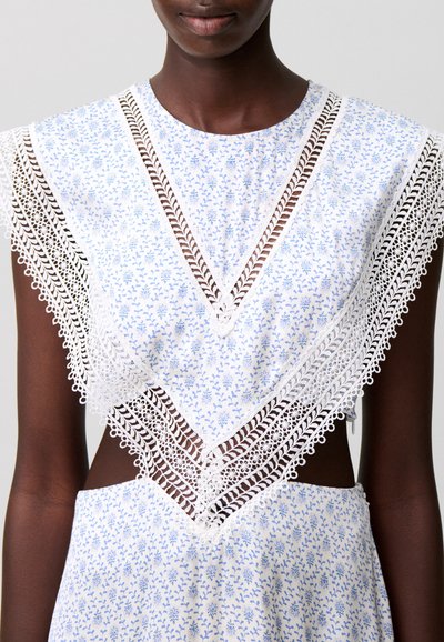 Donna che indossa un vestito bianco senza maniche con stampa floreale azzurro chiaro e dettagli intricati in pizzo bianco che formano una forma a V sul petto e sulle spalle.
