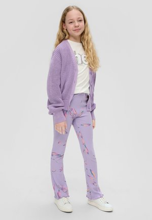 Lichtpaarse gebreide cardigan over een witte graphic T-shirt, gecombineerd met lichtpaarse broek met kleurrijke abstracte patronen, afgewerkt met witte sneakers.