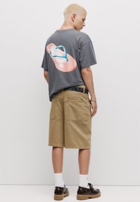 Grå t-shirt med et stort flip-flop grafik i pink og blå på bagsiden, parret med khaki shorts. Modellen har mørke sko og hvide sokker på.