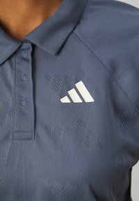 Námornícka modrá polokošeľa s textúrovaným vzorom, tri gombíky na golieri a biele logo Adidas na hrudi. Mäkký povrch látky.