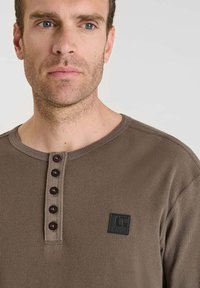 Hombre con una camisa henley de punto waffle marrón, con cinco botones y un pequeño parche rectangular negro en el pecho.