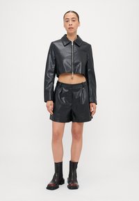Veste cropped en simili cuir noir avec fermeture éclair à l'avant, associée à un short assorti. Le mannequin porte des bottes chelsea noires avec une semelle épaisse.
