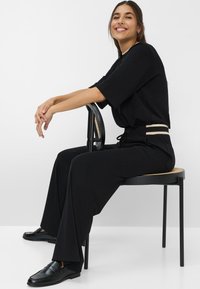 Schwarzes, locker sitzendes Outfit mit einem breiten Gürtel und schwarzen Schuhen, sitzt auf einem schwarzen Stuhl mit einer strukturierten Sitzfläche. Einfaches, sauberes Design, neutraler Hintergrund.